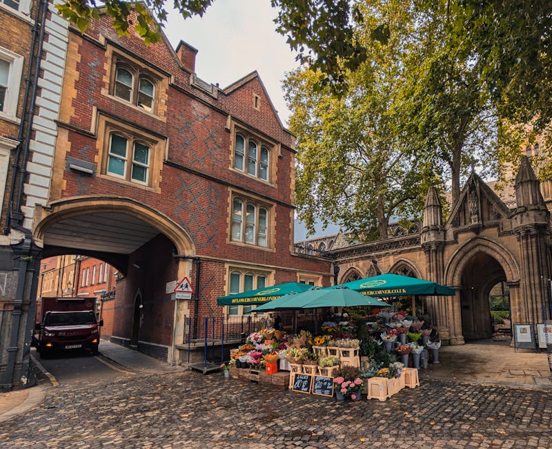 An outdoor flower stall located on a cobblestone street beneath an arched gateway, operated by Feltham Florist. The stall features an array of fresh, vibrant flowers including pink roses, yellow and p