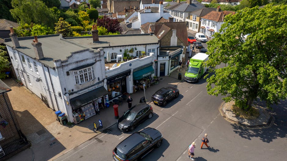 An aerial view of a small shopping street in Feltham, showing a white multi-story building with flower shops on the ground floor. The flower shop features a green awning and displays colorful floral a