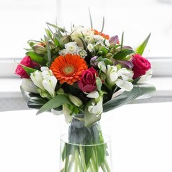 This captivating arrangement, prepared by florist artisans familiar with Feltham's vibrant community, is a masterful study in harmony and expression. At its heart lie large gerbera daisies in vivid orange and soft peach, their rounded petals forming a radiant halo that hints at the sunlit afternoons so common on Feltham Green. The deep magenta roses nestled within offer an intense, velvety contrast, their luxuriously swirled petals infusing the design with whispers of affection and admiration. Sprays of ivory-white alstroemeria and dainty white filler flowers create shimmering highlights that catch the light, bringing to mind the pristine blooms seen in local gardens after gentle rainfall. The whole ensemble is cradled by a lush collar of paddle-shaped, glossy aspidistra leaves, which curl protectively around the stems, grounding the arrangement and echoing the leafy tranquility of Feltham's tree-lined avenues. Finer greenery elegantly fills the spaces, creating texture and depth that captivates the senses. Each stem is placed with purpose and visible through the tall, slender glass vase-proof of a florist's skilled hand and dedication to quality. The bouquet sits in a softly illuminated setting, making the vibrant shades glow and the gentle perfume of roses and lilies linger in the air. Ideal for commemorating milestones, welcoming a new chapter, or simply brightening someone's day in Feltham, this bouquet reflects not just floral artistry, but the unique joy and optimism of the local area.